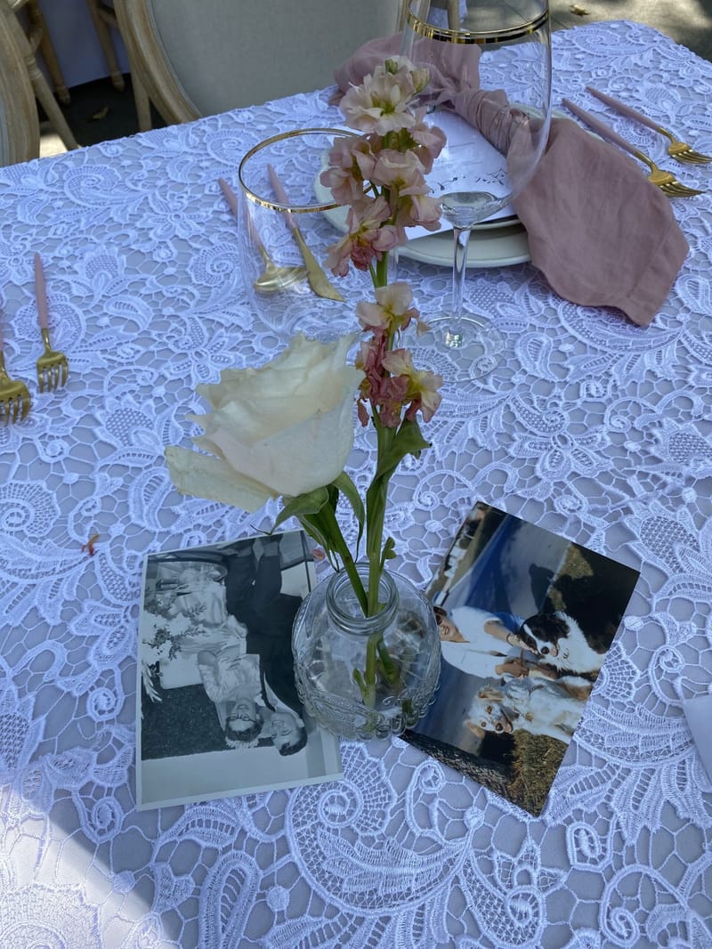 Old snapshots laid on a lace tablecloth around a bud vase with a white rose and pink stock