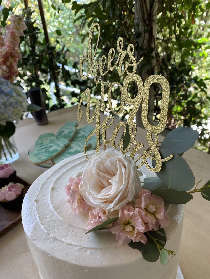 White frosted cake topped with a cream rose, pink stock and a gold glitter cheers to 90 years topper