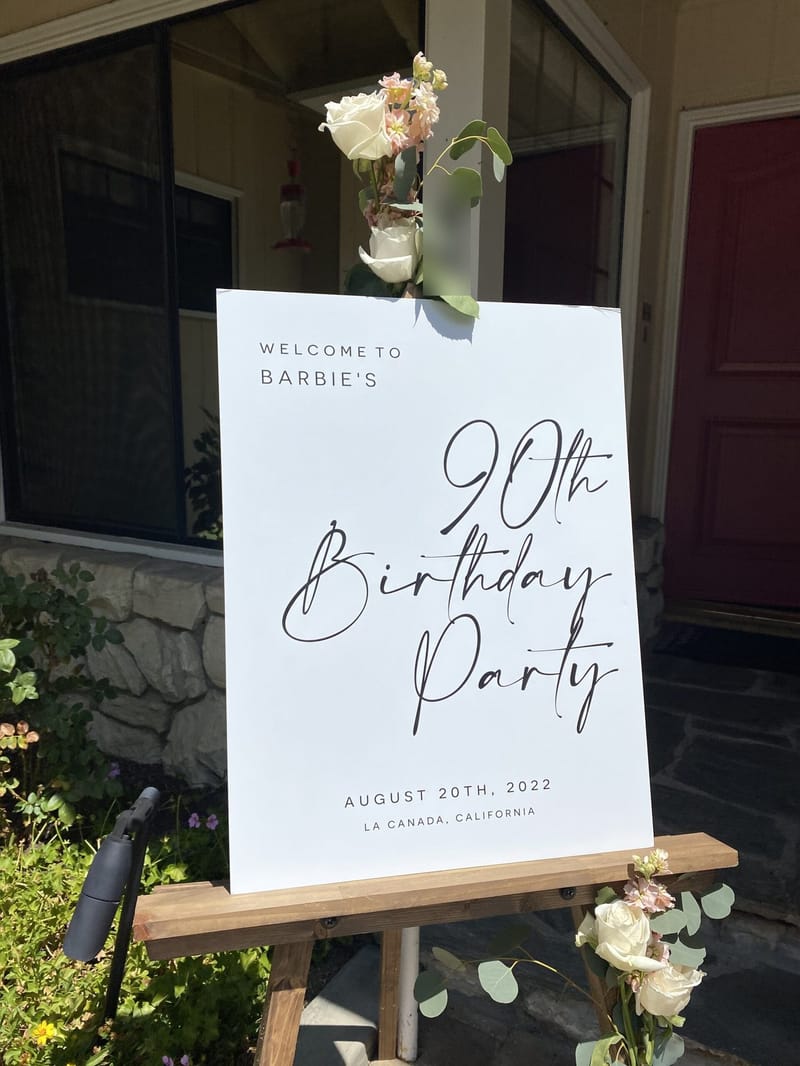 Welcome to Barbie’s 90th Birthday Party sign on a wooden easel beside a red front door