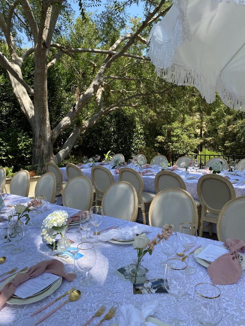 Lace-covered tables and cream oval-back chairs in a tree-shaded garden under a fringed white umbrella