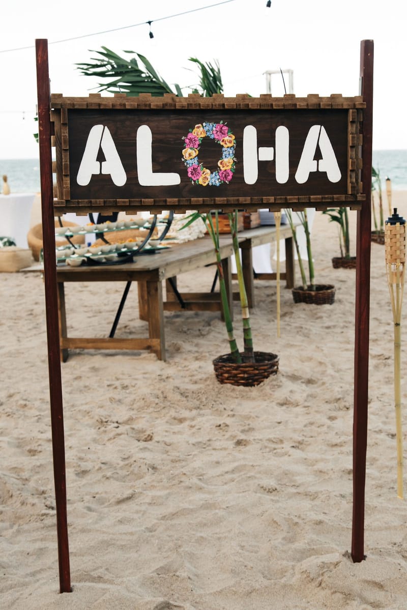 Wooden ALOHA sign with a ring of flowers for the O, standing in the sand before a buffet table