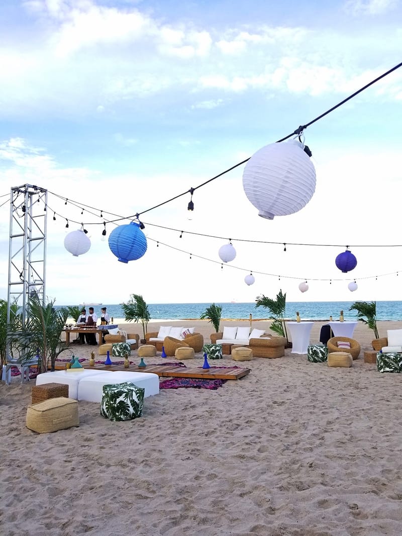 White and blue paper lanterns on string lights above lounge seating on the beach