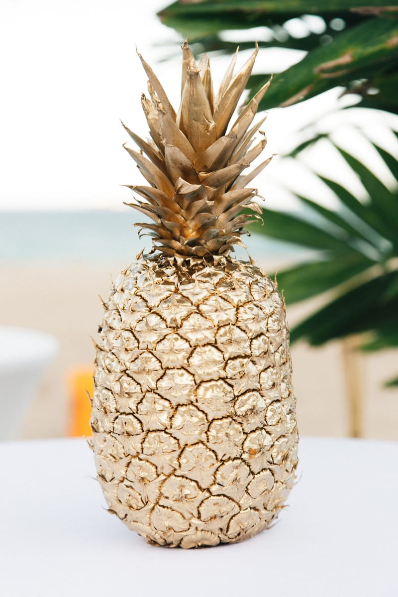 Gold-painted pineapple on a white table with palm leaves and the sea blurred behind
