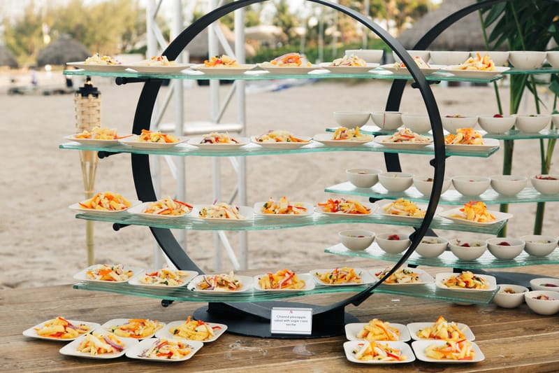 Glass stand on a black ring frame lined with small plates of charred pineapple salad on the sand