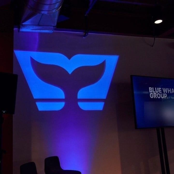 Whale tail logo projected in blue light on a wall beside a Blue Whale Group screen