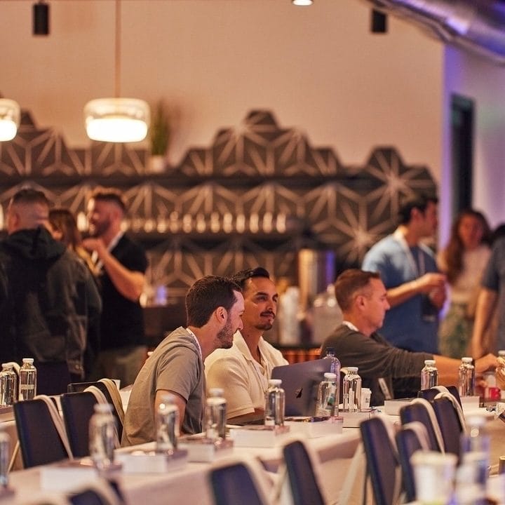 Attendees seated at long tables with water bottles in front of a geometric tiled wall