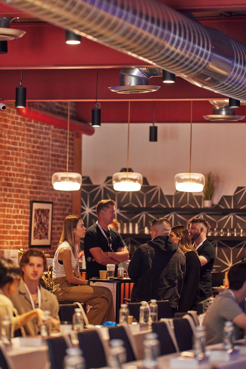 Attendees talking and seated at tables under pendant lights and exposed ductwork