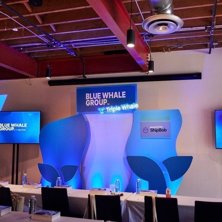 Blue wave-shaped stage backdrop with whale tail cutouts, a Blue Whale Group sign and a sponsored by ShipBob panel