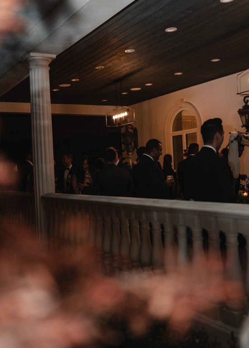 Guests in suits on a covered porch with white columns and a balustrade, pink flowers blurred in front