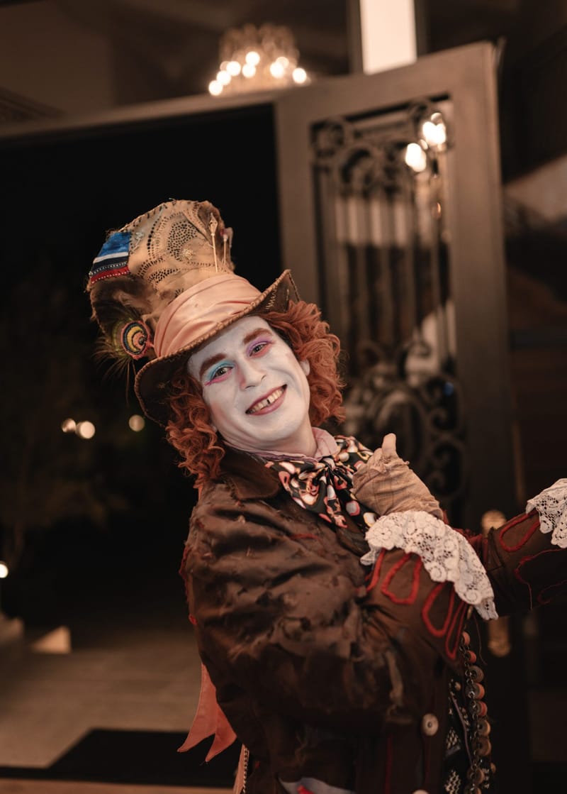 A smiling Mad Hatter in a patched top hat and lace cuffs beside an ornate iron door
