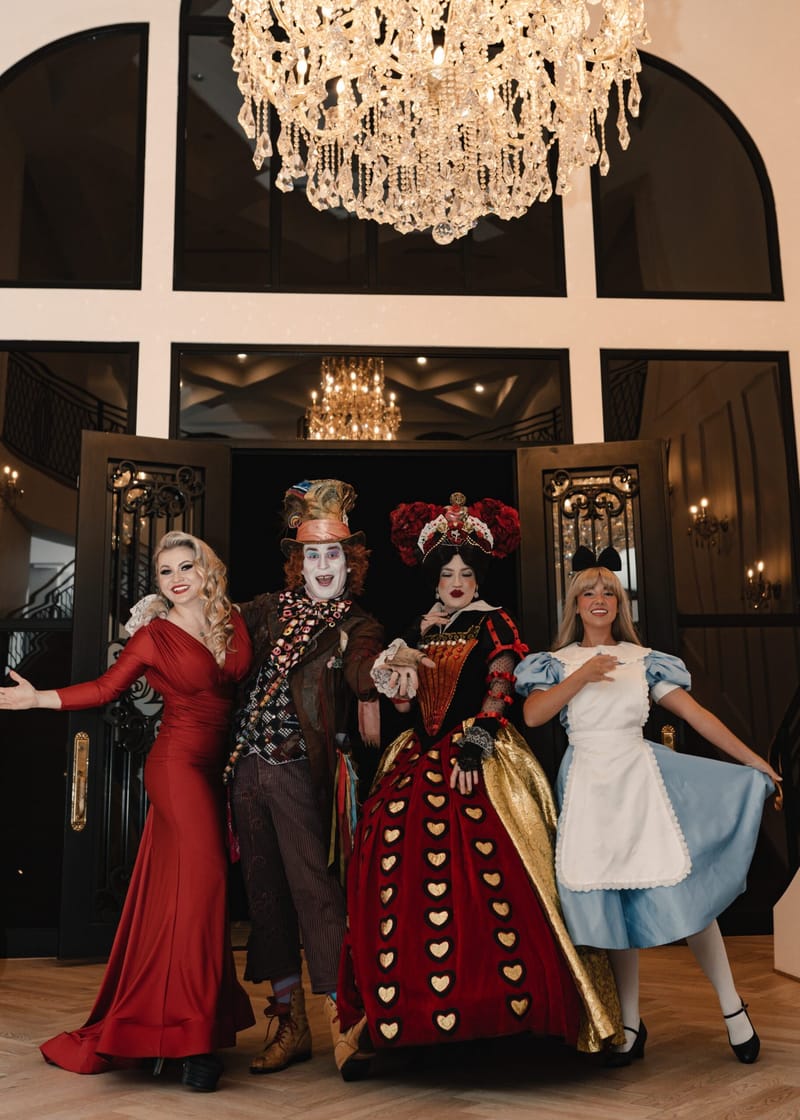A woman in a red gown, the Mad Hatter, the Queen of Hearts and Alice posing under a chandelier