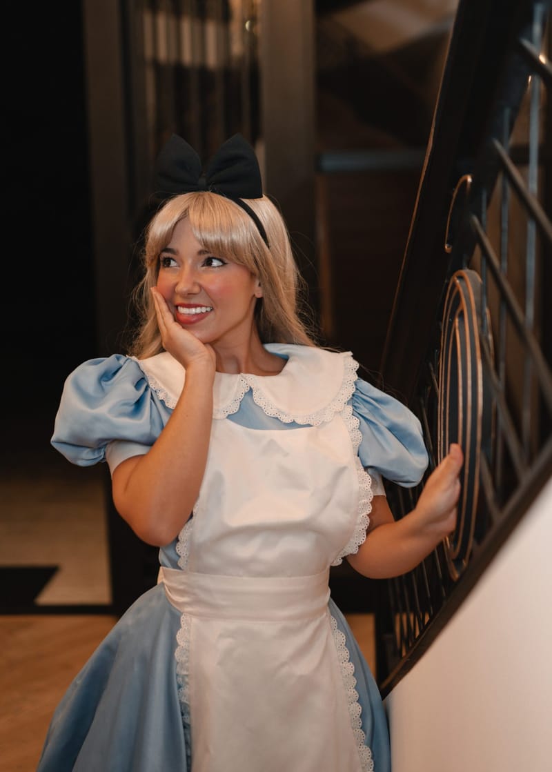 Alice in a blue dress and white apron posing on a staircase