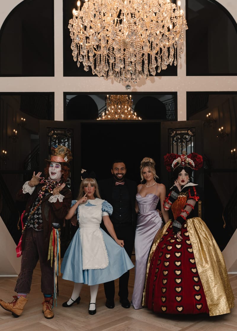 The Mad Hatter, Alice, the Queen of Hearts and two guests posing under a crystal chandelier