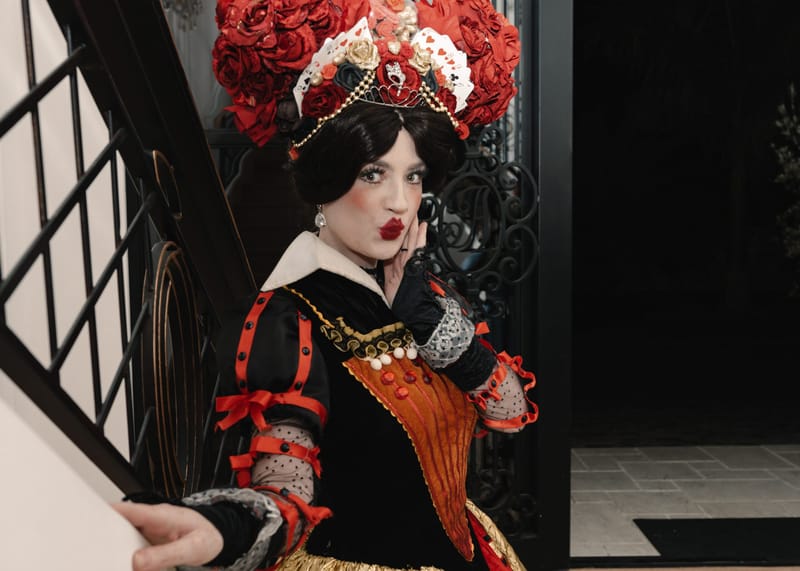 The Queen of Hearts in a rose headdress blowing a kiss at the foot of a staircase