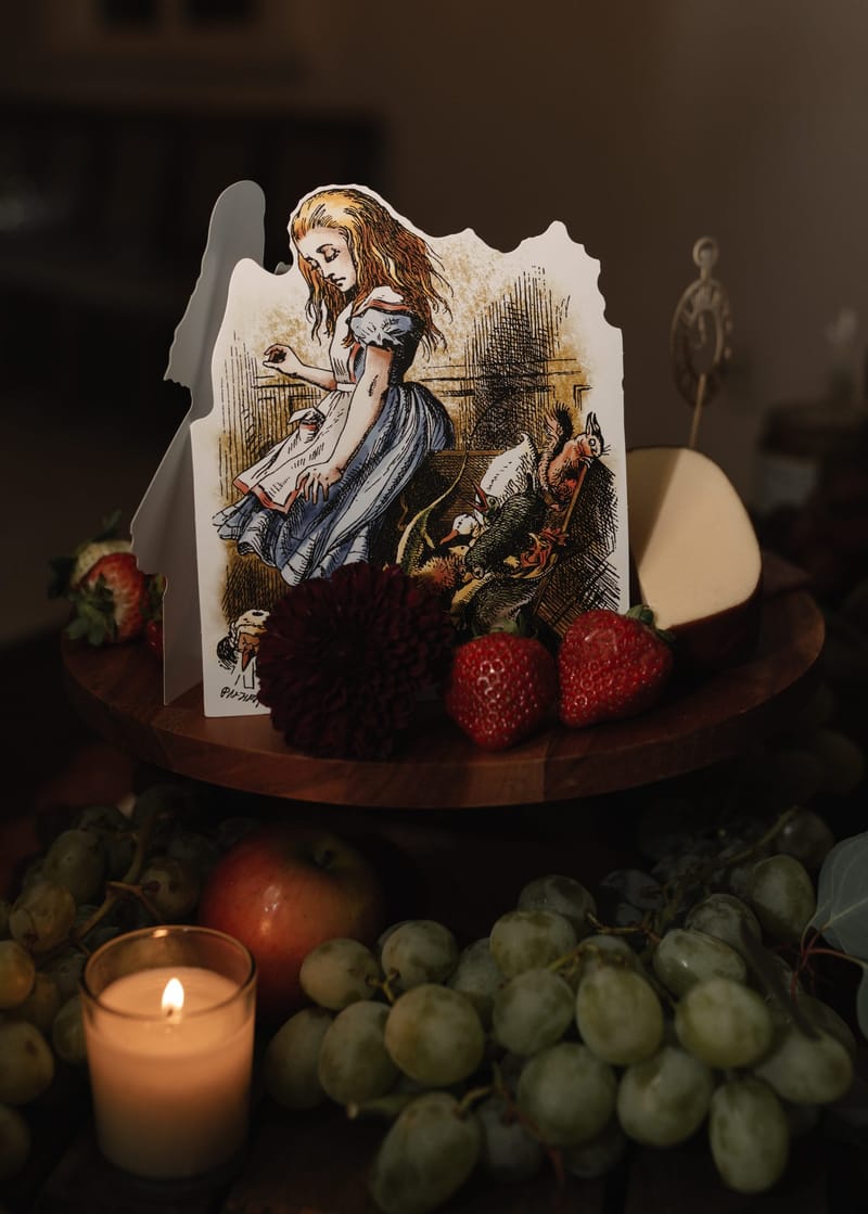 Illustrated Alice cutout on a wooden board with strawberries, a dahlia, grapes and a candle