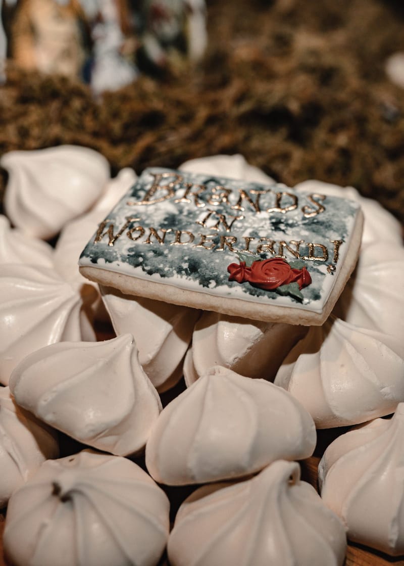 Cookie lettered in gold with Brands in Wonderland! and a red icing rose, resting on meringues
