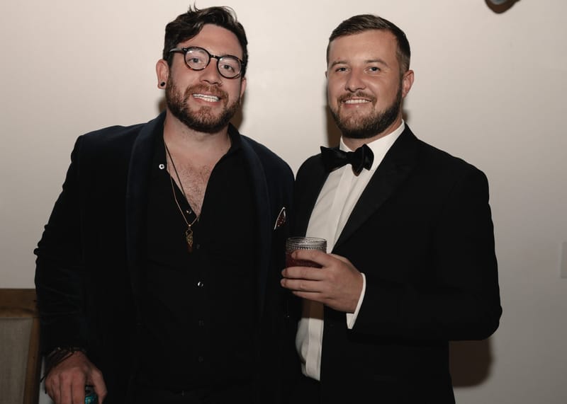Two guests in black jackets smiling for the camera, one holding a drink
