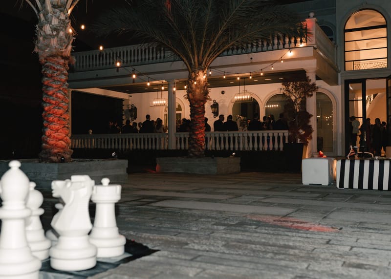 Giant white chess pieces in a courtyard with palm trees and string lights outside a lit terrace