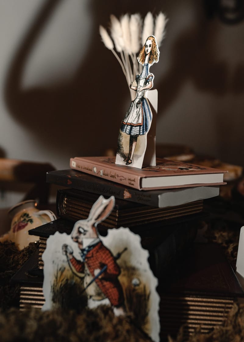 Illustrated Alice and White Rabbit cutouts on a stack of books with pampas grass