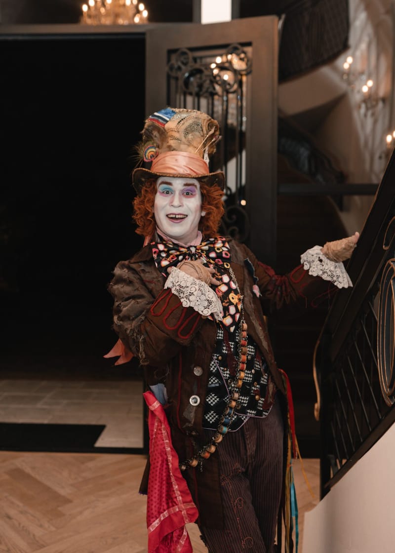 The Mad Hatter gesturing from a staircase beside an open iron door