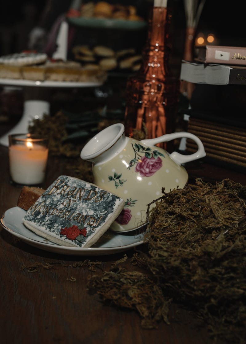 Floral teacup and saucer with a Brands in Wonderland cookie among moss beside a candle