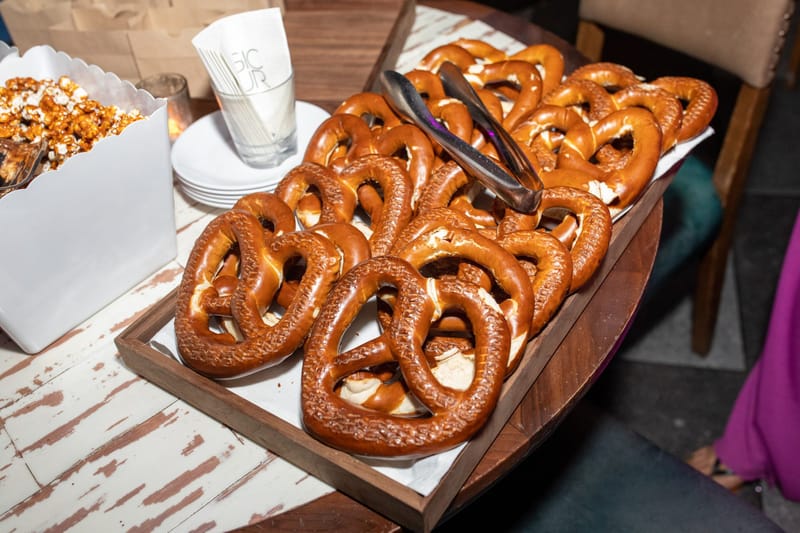 Tray of soft pretzels with tongs beside a box of caramel popcorn on a whitewashed table