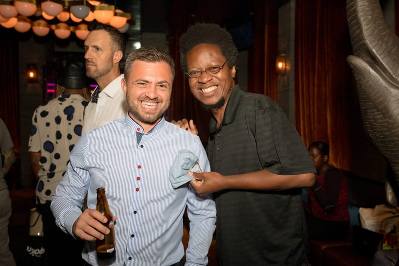 Two smiling guests, one holding a beer bottle, in a lounge with velvet curtains