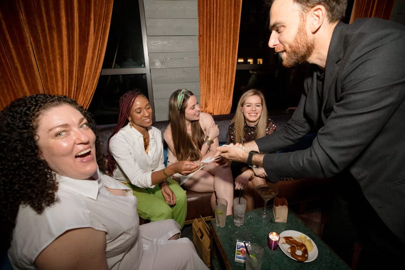 A man in black leaning in to show something in his hands to laughing guests on a sofa