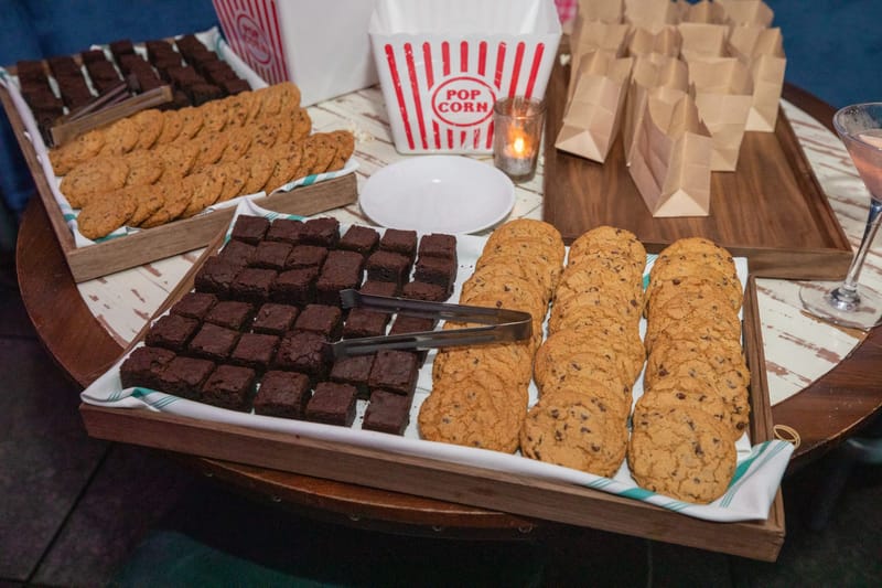 Trays of chocolate chip cookies and brownies beside a striped popcorn box and paper bags