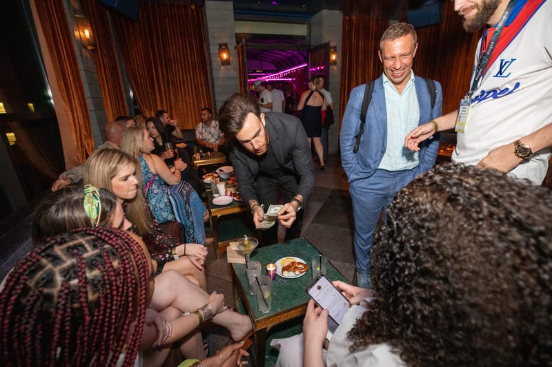 A man in black holding playing cards over a low table as guests watch in the lounge