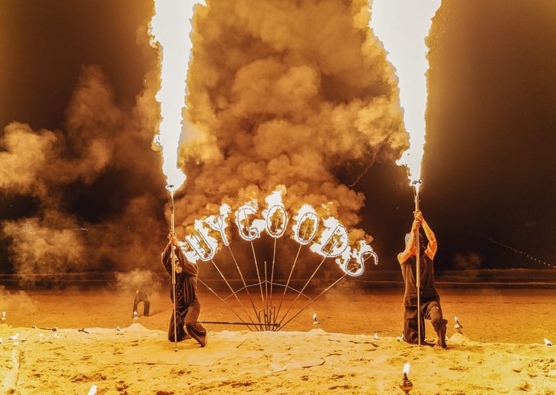 Two fire performers holding tall flame jets beside a burning BUYGOODS sign on the sand