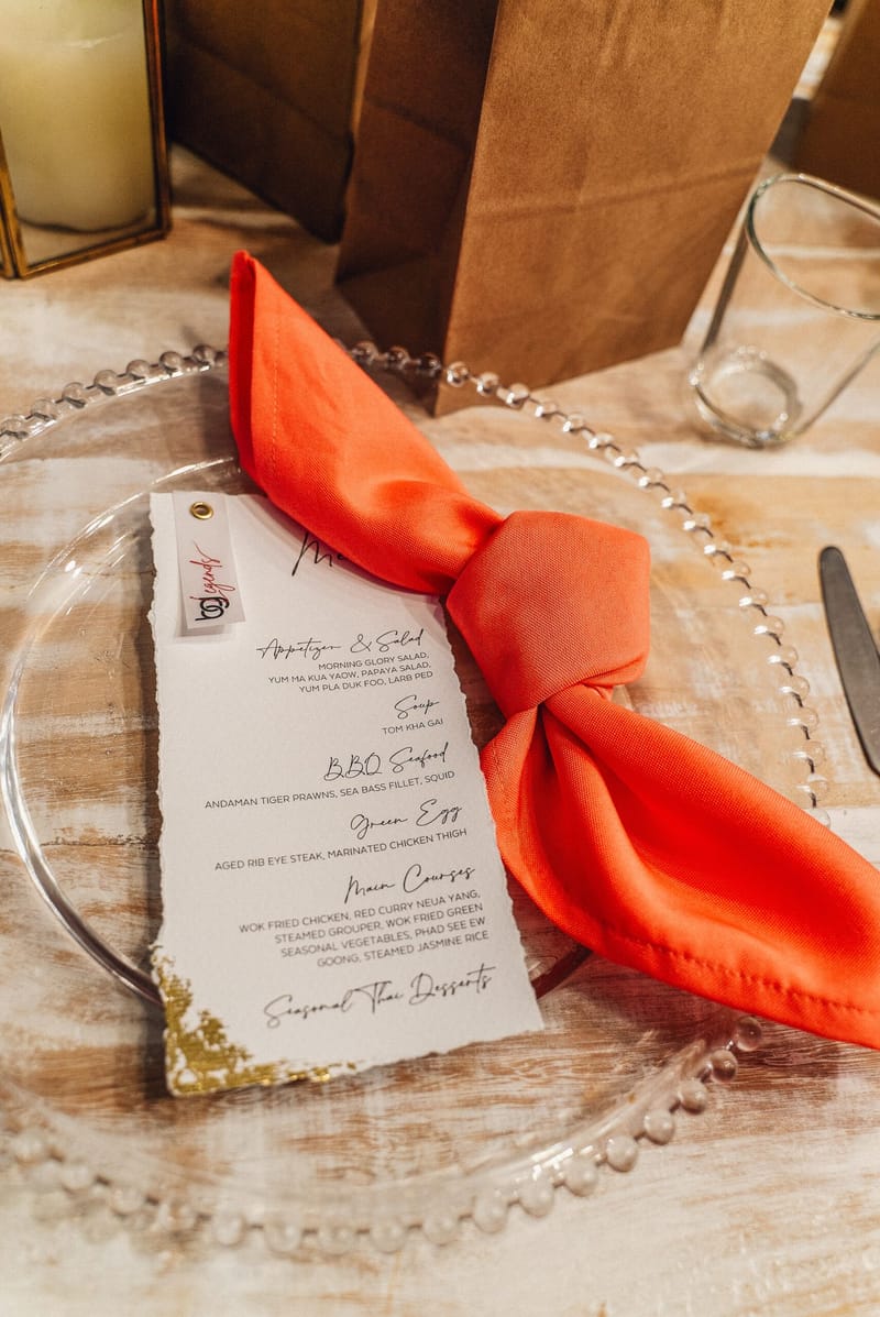 Thai dinner menu card with a small logo tag beside a knotted coral napkin on a beaded glass charger