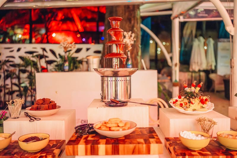 Chocolate fountain on white plinths with dipping bowls, a pineapple fruit platter and wooden boards