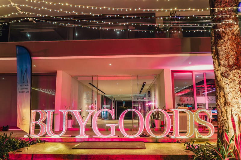 Illuminated BUYGOODS marquee letters in front of a pink-lit building under string lights