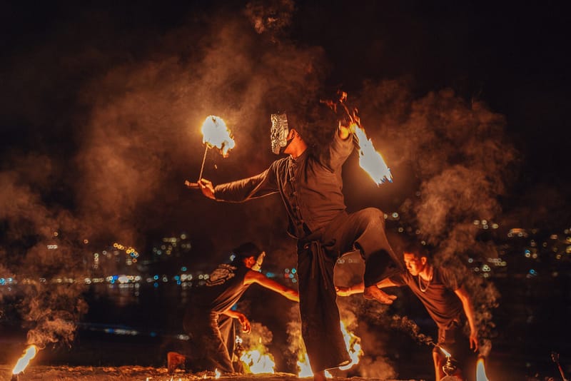 Masked fire performers spinning flaming props on the beach at night, city lights across the water