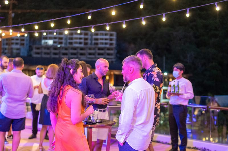 Guests mingling on a terrace under string lights as a masked server carries drinks