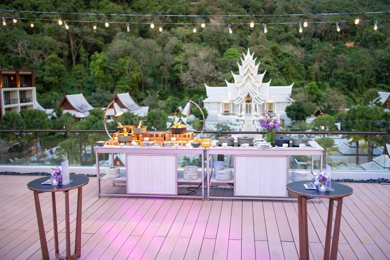 Buffet stations on a deck under string lights, with a white temple-style building on the hillside