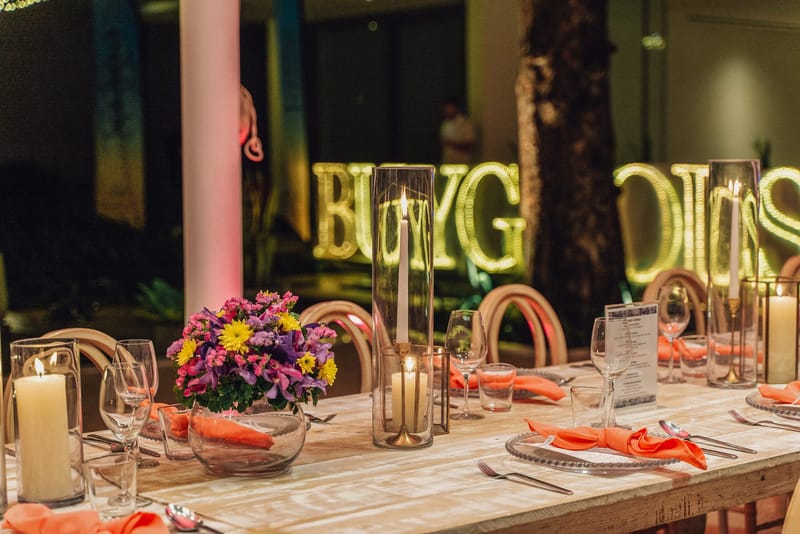 Long dinner table with candles in glass cylinders, flowers and coral napkins before lit BUYGOODS letters
