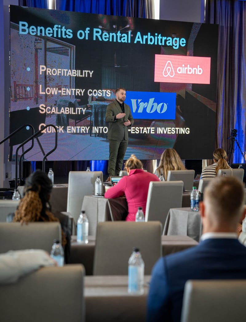 Speaker presenting Benefits of Rental Arbitrage, with Airbnb and Vrbo logos, to guests at draped tables