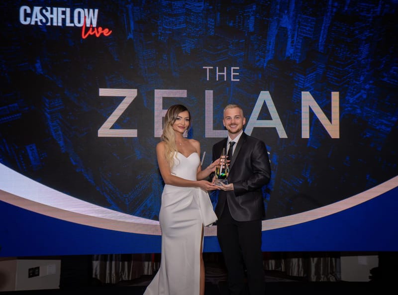 A woman and a man holding a glass trophy in front of a Cashflow Live screen reading The Zelan