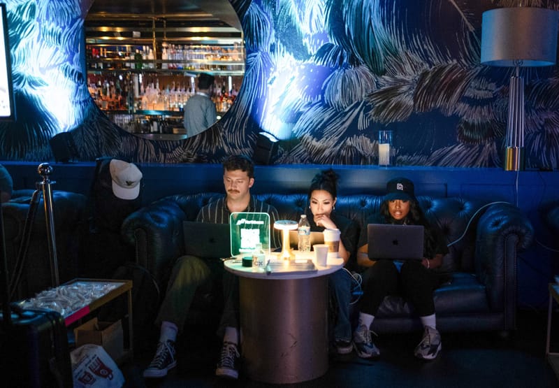 Three guests working on laptops in a blue tufted leather banquette beside a lit Northbeam QR sign