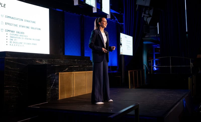A speaker in a dark suit on stage beside a slide listing communication structure and company values