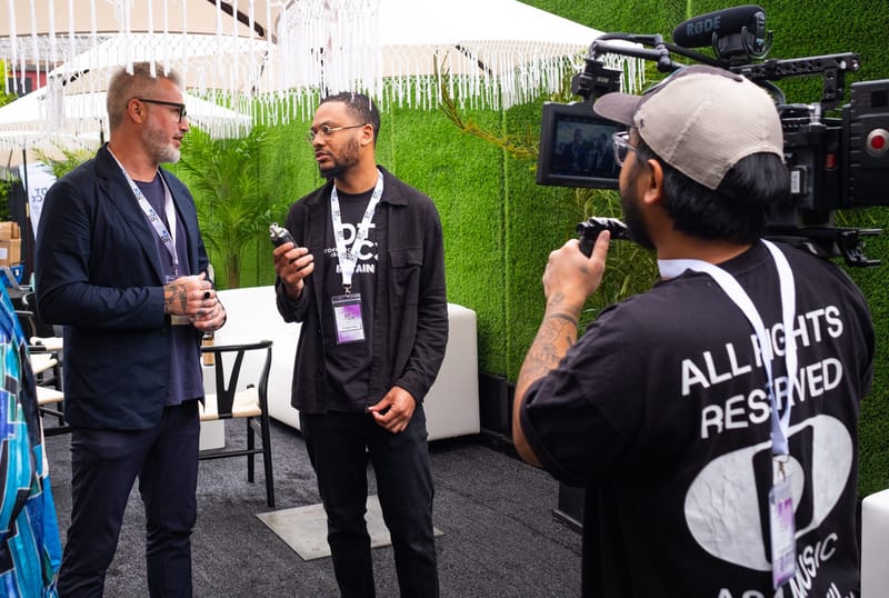 Two attendees in DTCx lanyards filmed by a camera operator in front of a green hedge wall