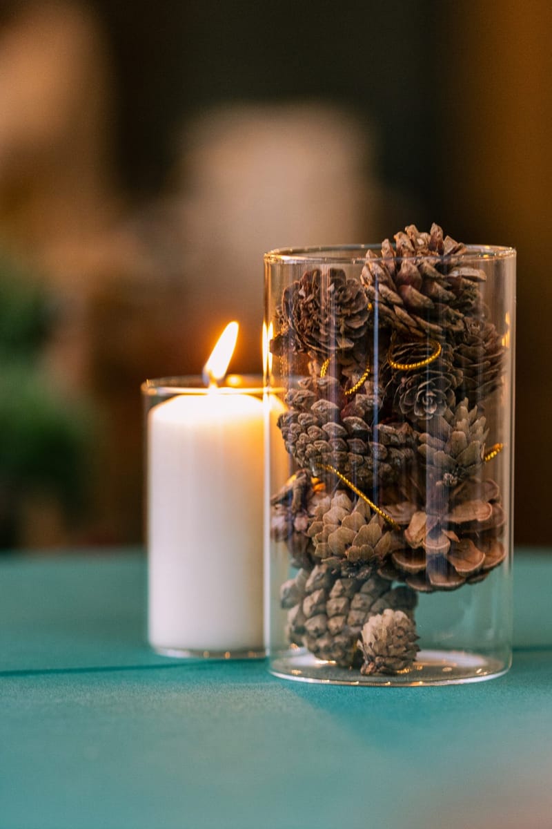 Glass cylinder filled with pinecones beside a lit pillar candle on a green table