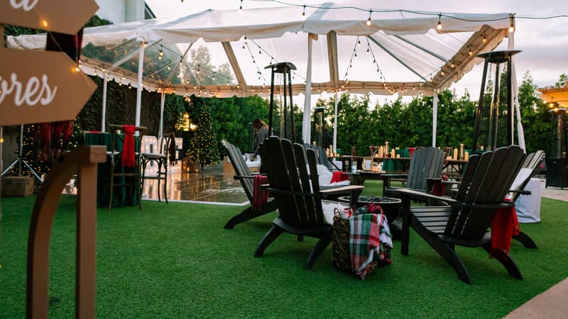 Adirondack chairs and a plaid blanket on artificial grass beside a clear tent with patio heaters