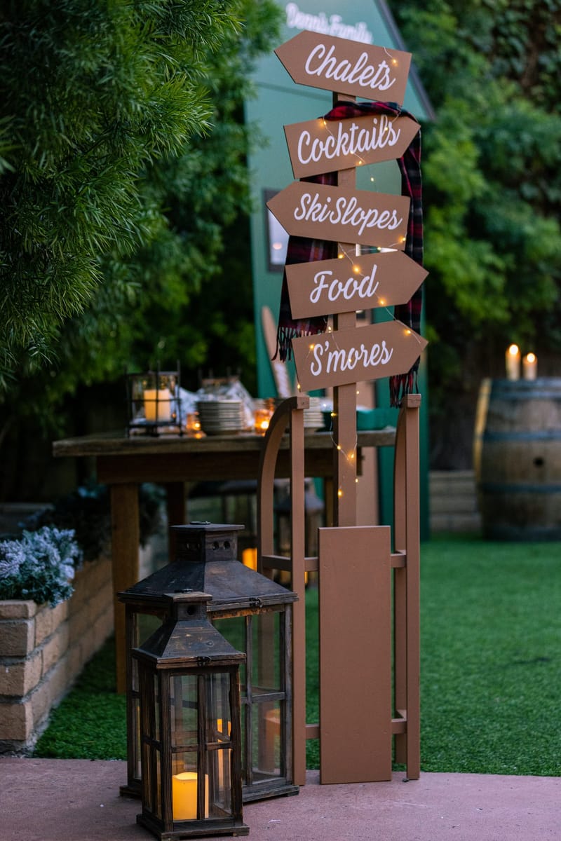 Wooden signpost and lanterns on a path beside a green cabin and a wine barrel