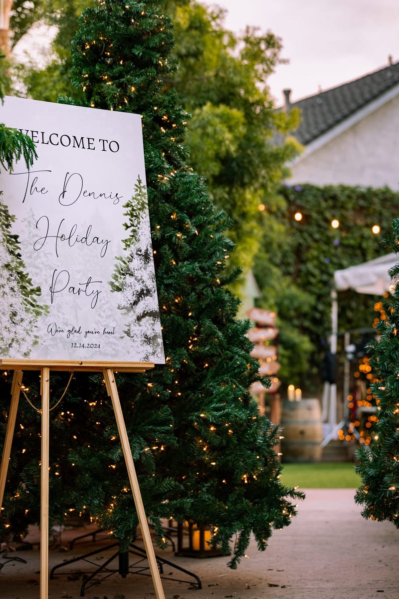 Welcome to the Dennis Holiday Party sign on an easel beside a lit Christmas tree