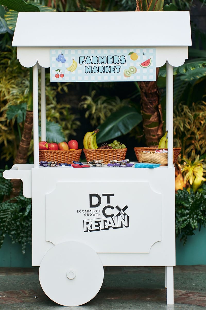 White Farmers Market cart with fruit baskets and snacks, printed with the DTCx Retain logo