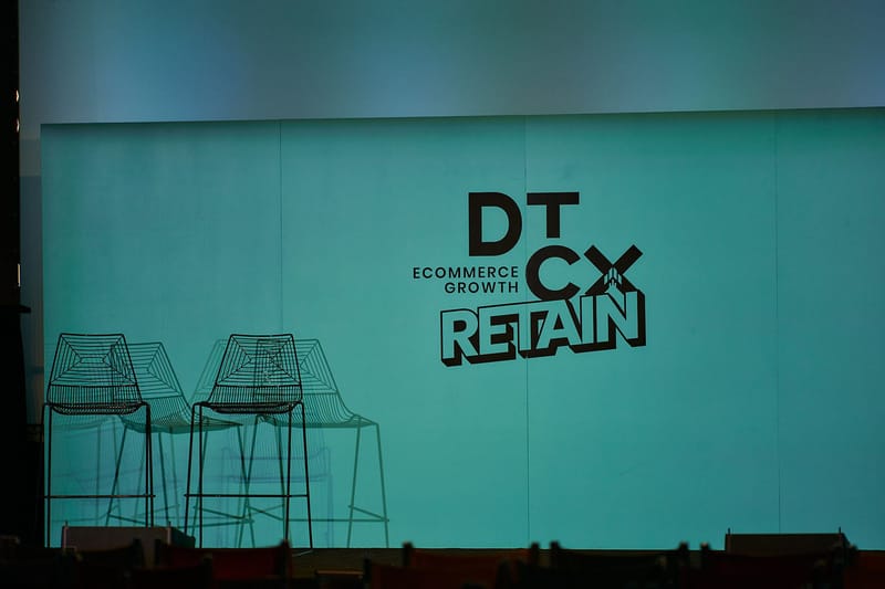 Teal stage backdrop printed with the DT CX Ecommerce Growth Retain logo, with two wire stools