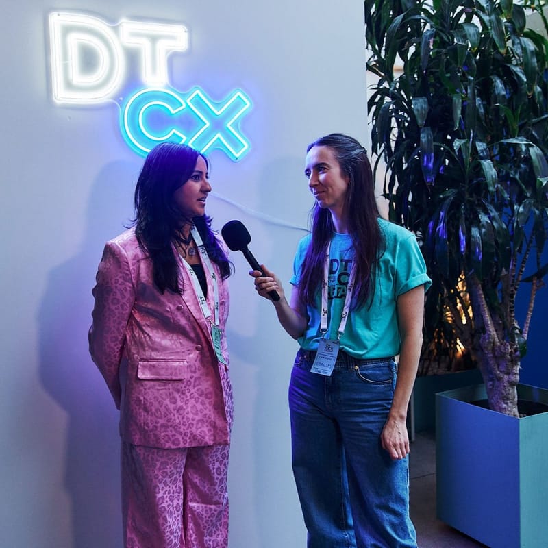 One guest interviewing another with a microphone under a neon DT CX sign